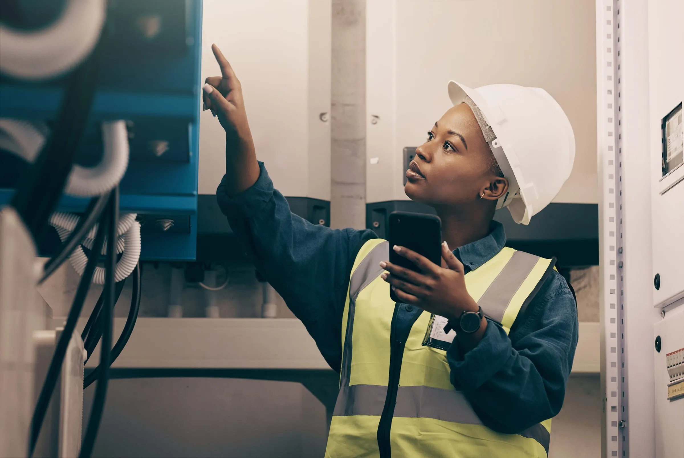 A technician in a hard hat and safety vest checks her phone in an electrical room.