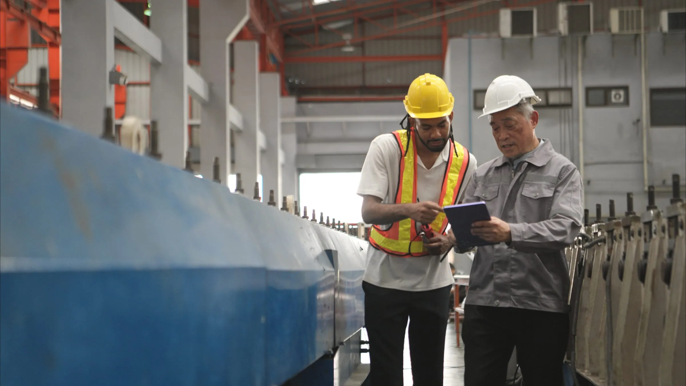 Two maintenance engineers in hard hats review a production line with a tablet.